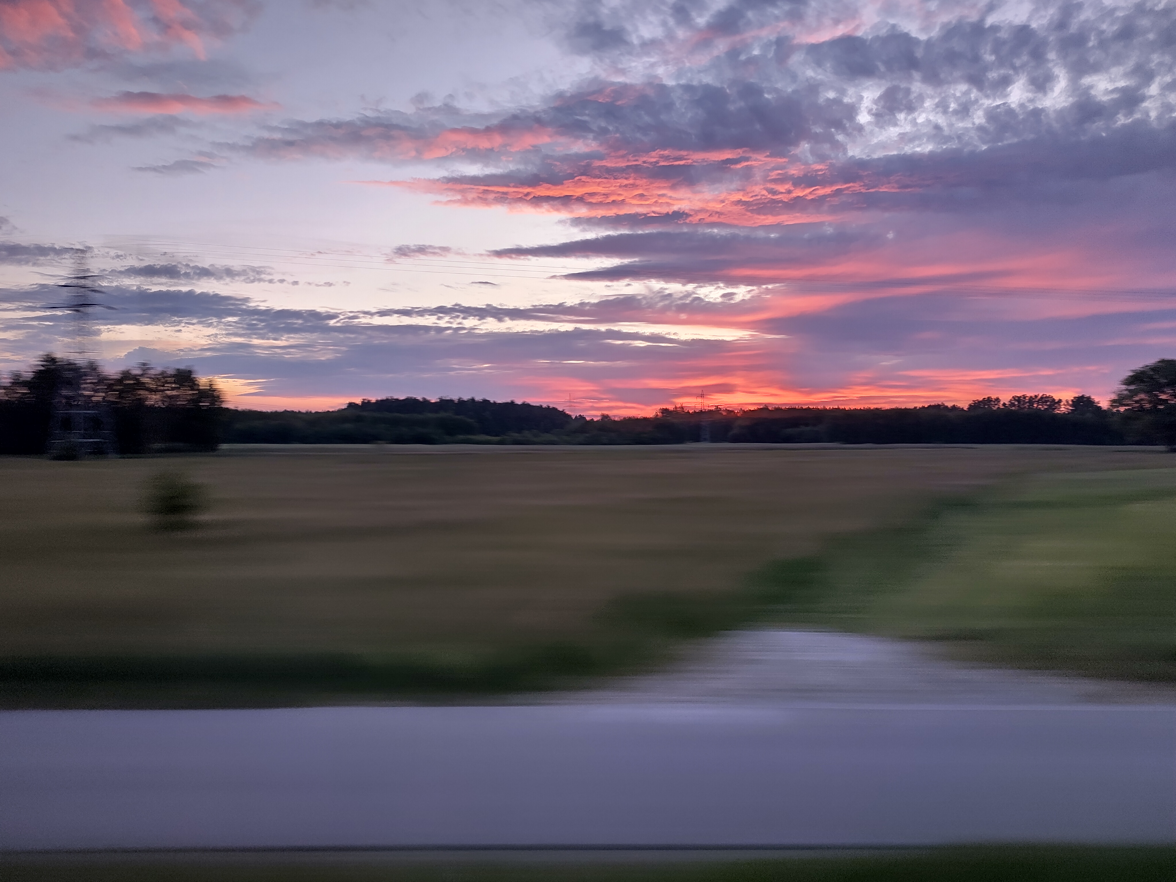 RUSH!: it&rsquo;s a picture of a sunset above a green flatland in Poland. In foreground we see mainly grass, a small road and a couple of transmission towers. Further away some trees hiding the horizon and the sun from our sight. This picture was taken from a moving train, which means that everything below the horizon looks blurry as it&rsquo;s blazing past the viewer and the camera, whereas the sky and clouds look still, showing how far they avtually are from us.