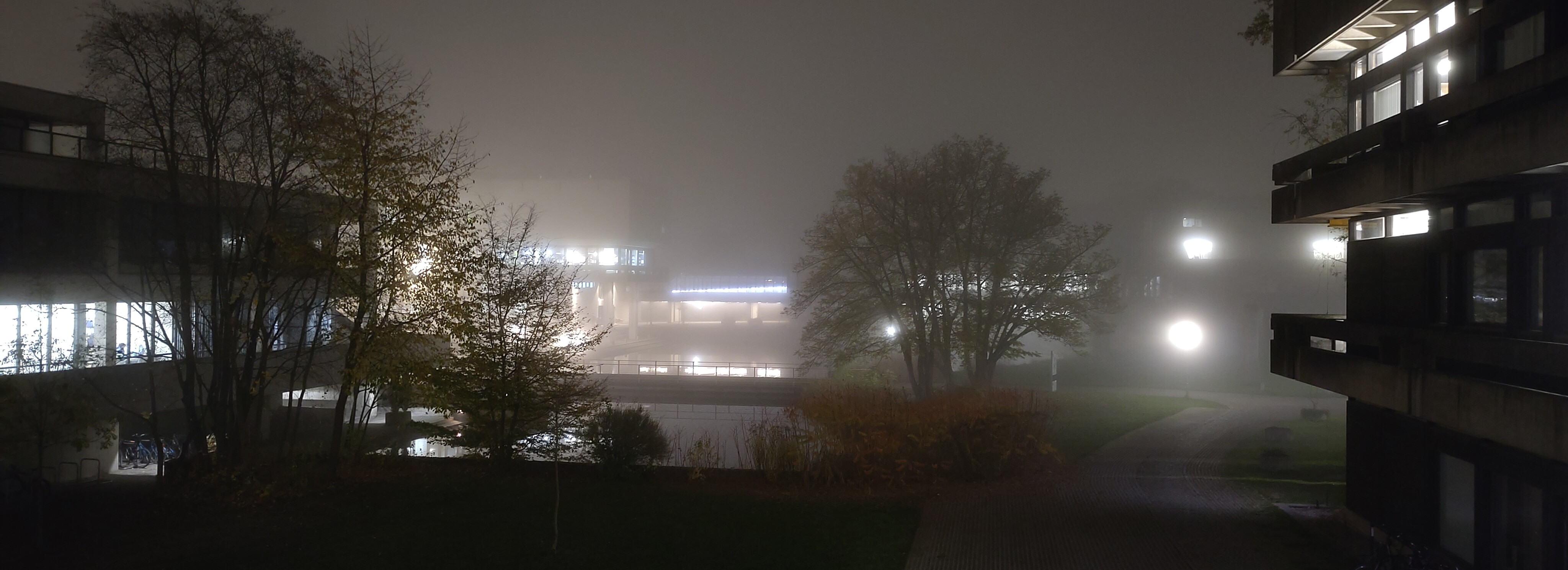 a picture of a very well lit building, standing in front of a small lake with a bridge cutting through it. It's a foggy night, so the background is mainly visible through the lights, and the volumes of the building cannot be easily distinguished.
